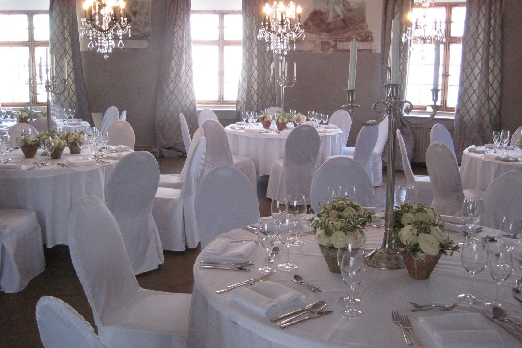 Hoher Saal mit hohen, lichten Fenstern und Kronleuchtern dazwischen; Runde, weiss eingedeckte Tische mit glitzernden Besteck und Gläsern und Blumenbouquet